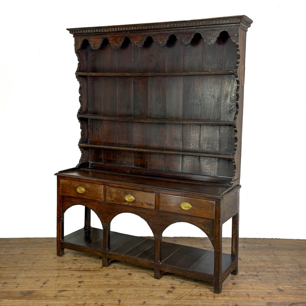 A dark oak 19th-century Welsh dresser featuring a three-tier open plate rack with a scalloped canopy above three drawers and an open pot board lower section. View IX