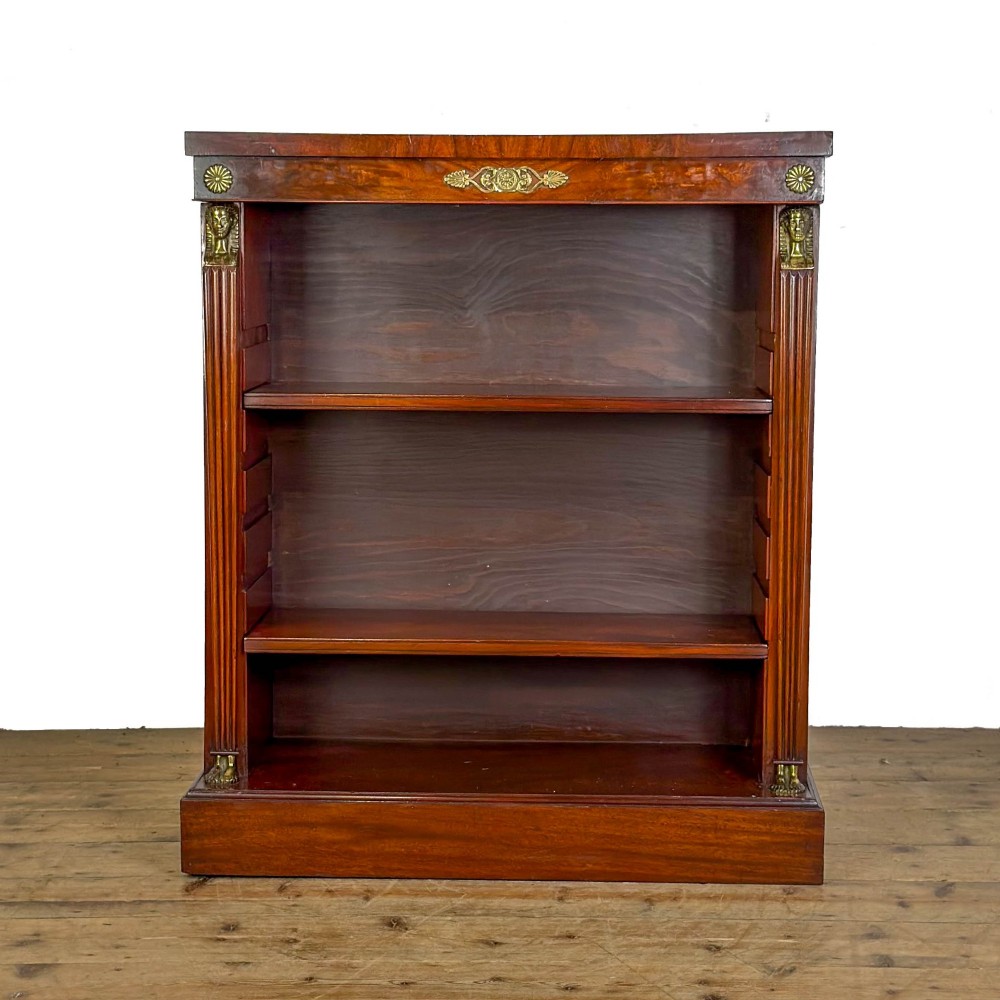 An antique Egyptian Revival mahogany open bookcase featuring fluted pilasters topped with cast brass sphinx heads, adjustable wooden shelves, and a plinth base.