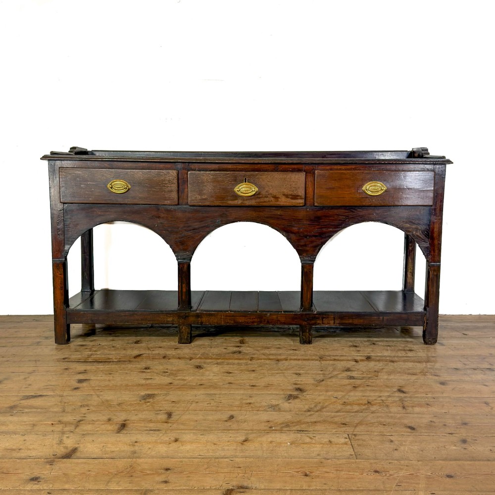 A 19th-century dark oak dresser base featuring three upper drawers with brass oval handles, three decorative arched aprons, and a low potboard shelf.