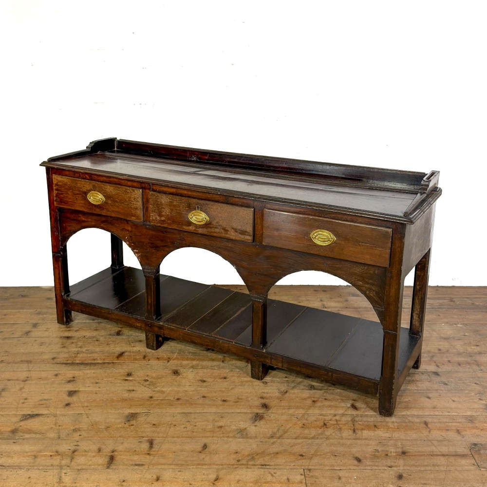 A 19th-century dark oak dresser base featuring three upper drawers with brass oval handles, three decorative arched aprons, and a low potboard shelf. View VII