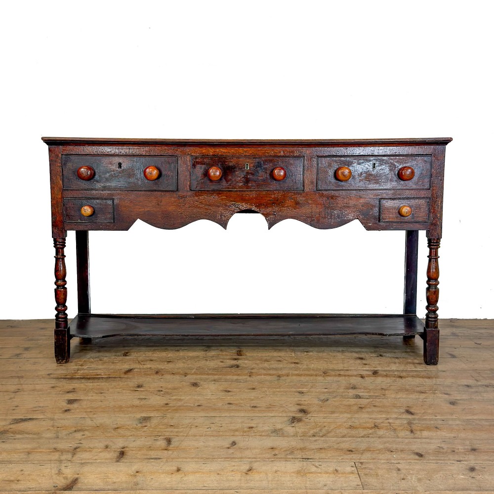 An antique dark wood dresser base featuring turned front legs, a scalloped apron, multiple drawers with wooden knob handles, and a lower pot board shelf.