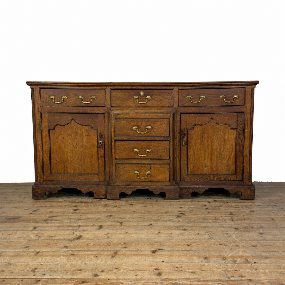 An antique Georgian oak dresser base with multiple drawers, fielded panel cupboard doors, and brass swan-neck handles on a bracket feet base.