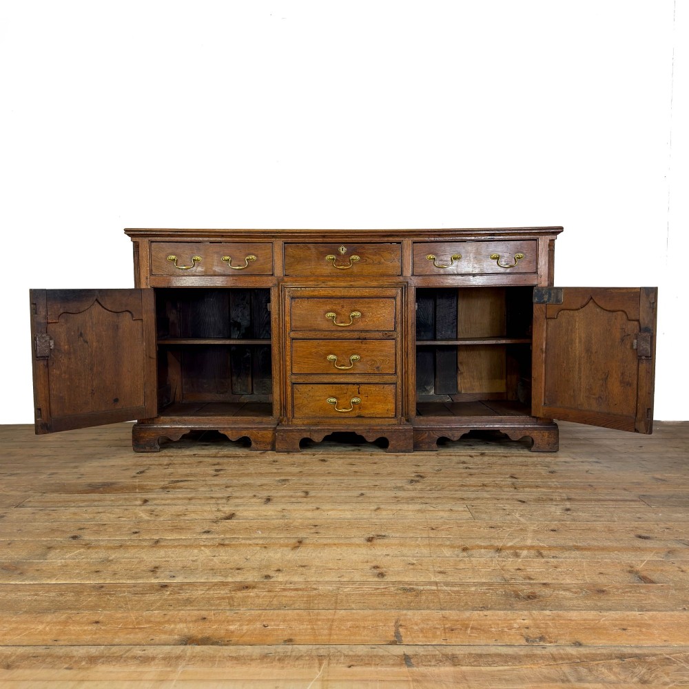 An antique Georgian oak dresser base with multiple drawers, fielded panel cupboard doors, and brass swan-neck handles on a bracket feet base. View II