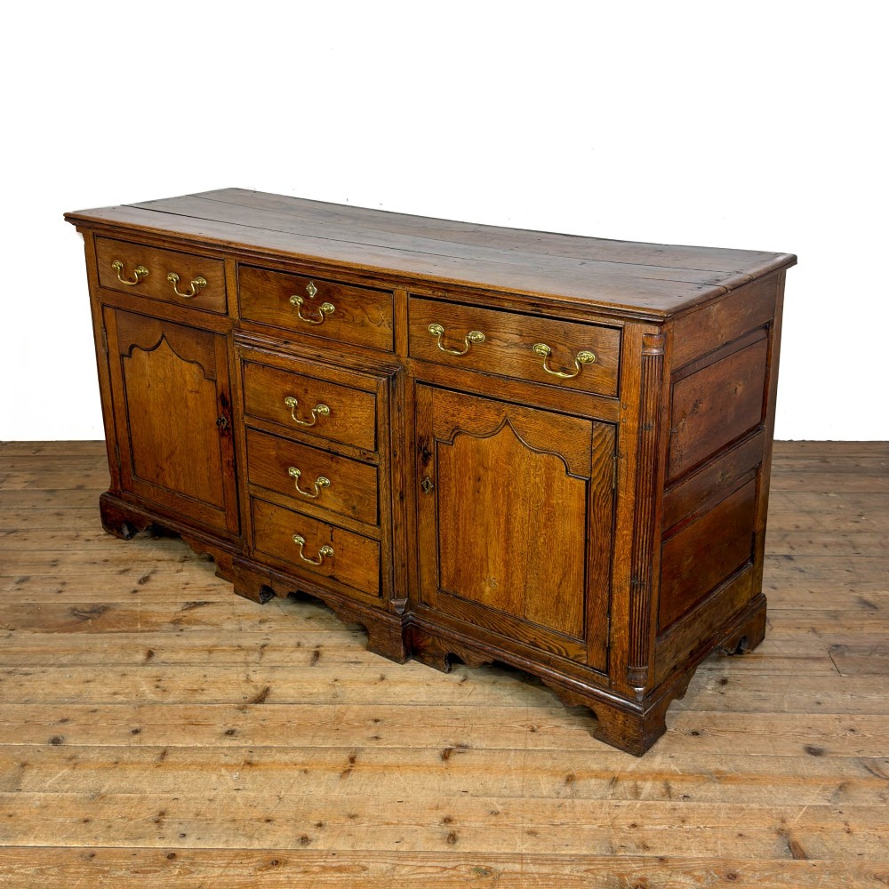 An antique Georgian oak dresser base with multiple drawers, fielded panel cupboard doors, and brass swan-neck handles on a bracket feet base. View V