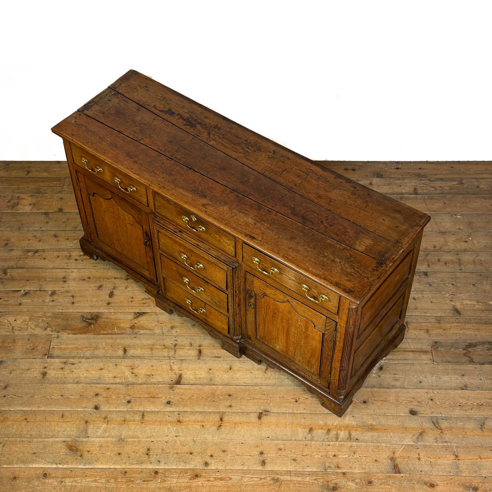 An antique Georgian oak dresser base with multiple drawers, fielded panel cupboard doors, and brass swan-neck handles on a bracket feet base. View VI