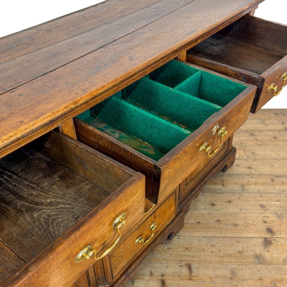 An antique Georgian oak dresser base with multiple drawers, fielded panel cupboard doors, and brass swan-neck handles on a bracket feet base. View XIV