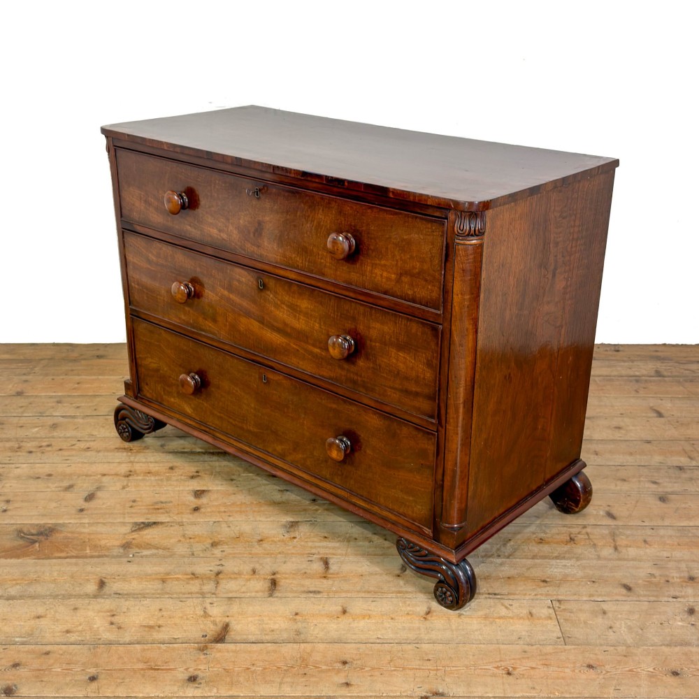 A William IV dark mahogany chest of drawers featuring three long drawers with turned knobs, brass escutcheons, and carved scroll feet. View II