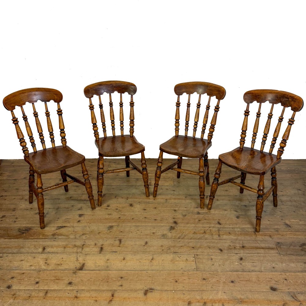 A matching set of four wooden farmhouse kitchen chairs featuring turned spindle backs, contoured seats, and turned legs connected by H-stretchers.