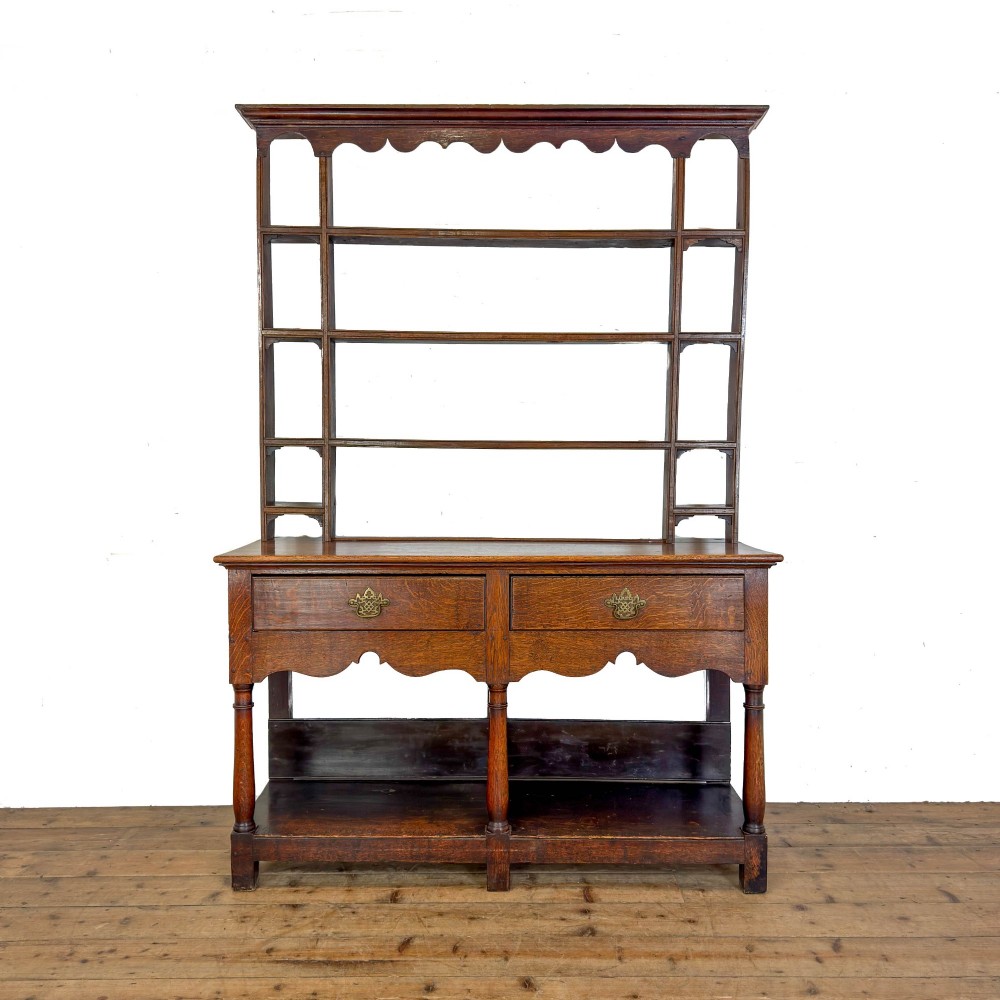 An antique oak Welsh dresser featuring an open tiered plate rack with a scalloped top frieze over two drawers and a bottom pot board shelf.