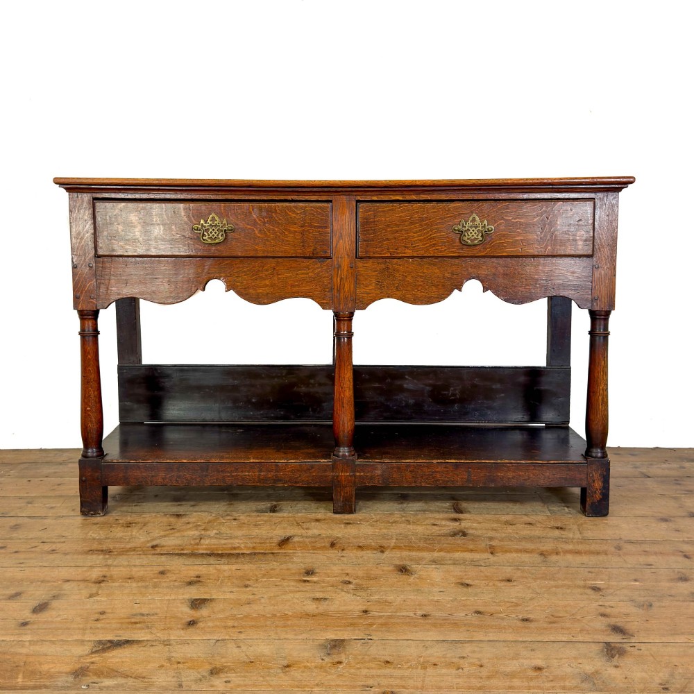 An antique oak dresser base featuring two drawers with brass backplate handles, shaped lower aprons, turned front legs, and an open potboard shelf.