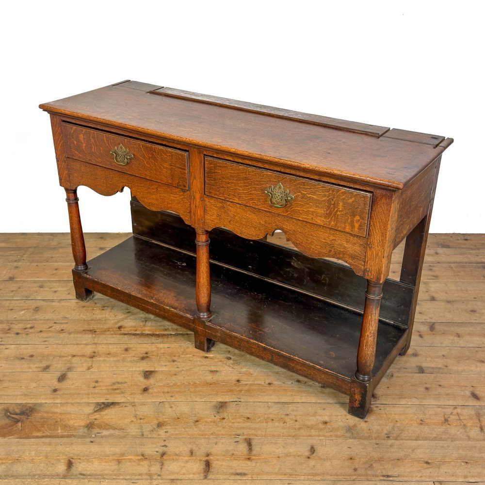 An antique oak dresser base featuring two drawers with brass backplate handles, shaped lower aprons, turned front legs, and an open potboard shelf. View III