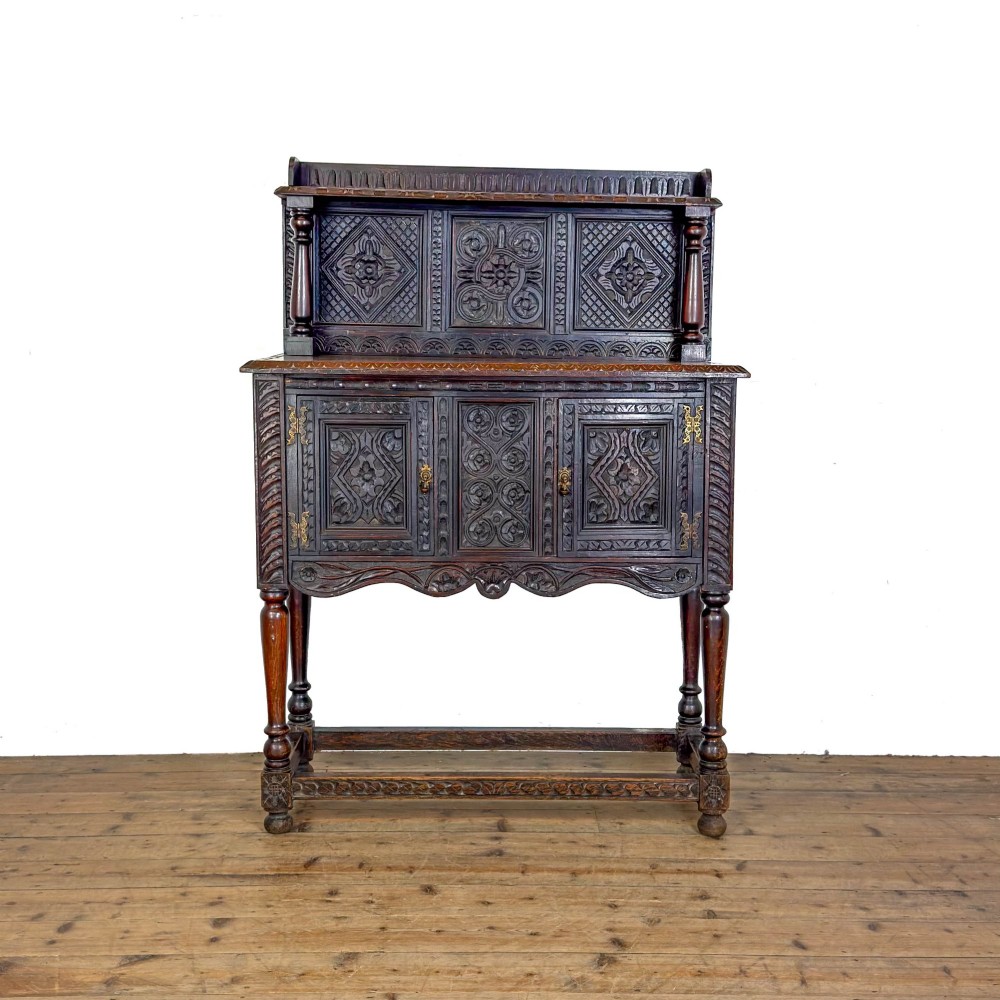 A dark carved oak Victorian sideboard with a tiered top shelf, detailed geometric carved door panels, brass hinges, and turned legs with a lower stretcher rail.