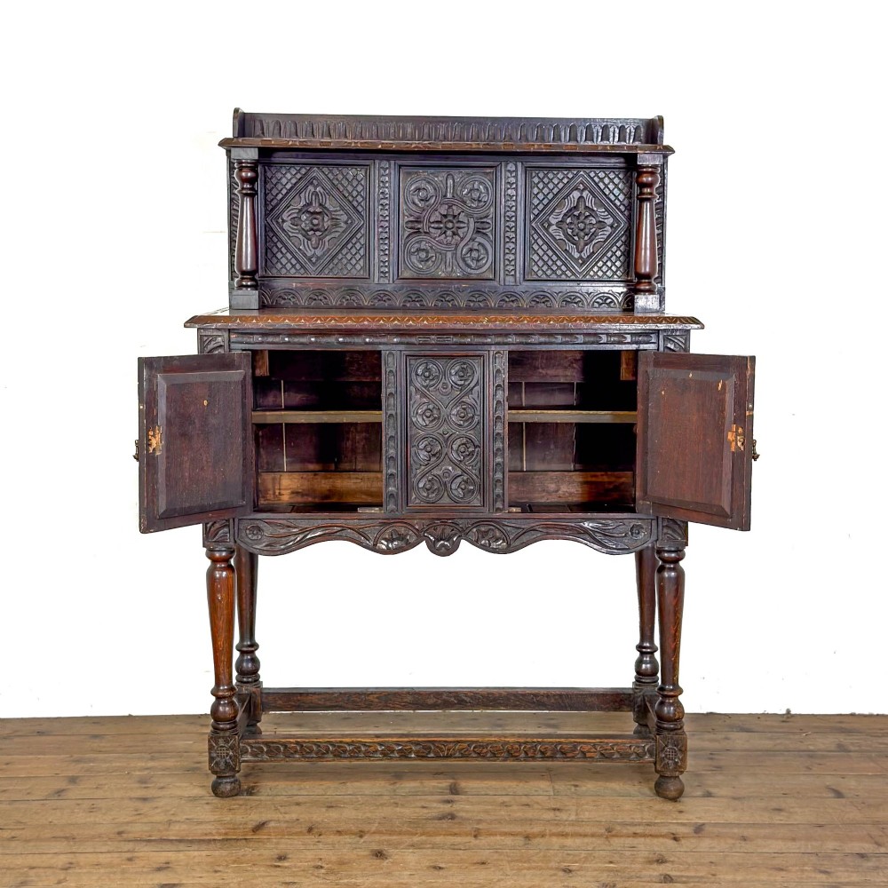 A dark carved oak Victorian sideboard with a tiered top shelf, detailed geometric carved door panels, brass hinges, and turned legs with a lower stretcher rail. View II