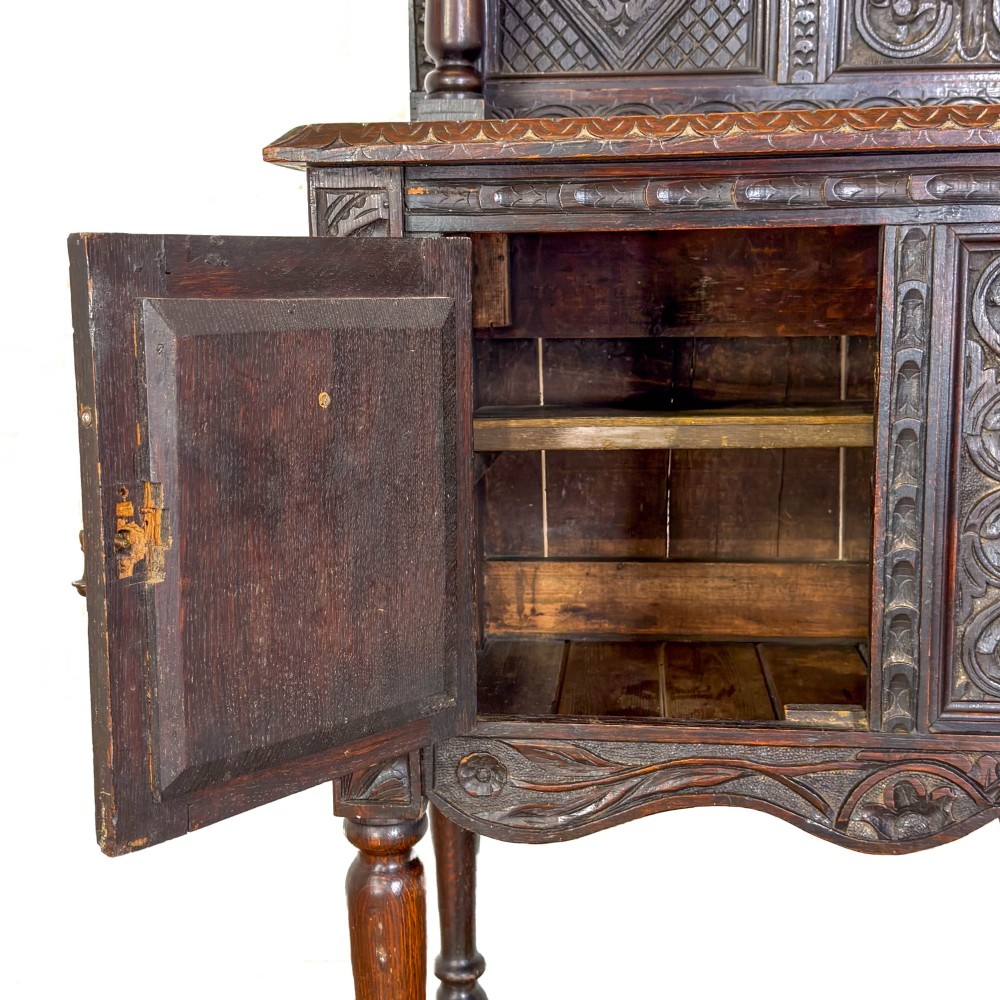A dark carved oak Victorian sideboard with a tiered top shelf, detailed geometric carved door panels, brass hinges, and turned legs with a lower stretcher rail. View III