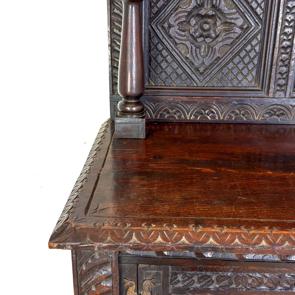 A dark carved oak Victorian sideboard with a tiered top shelf, detailed geometric carved door panels, brass hinges, and turned legs with a lower stretcher rail. View V