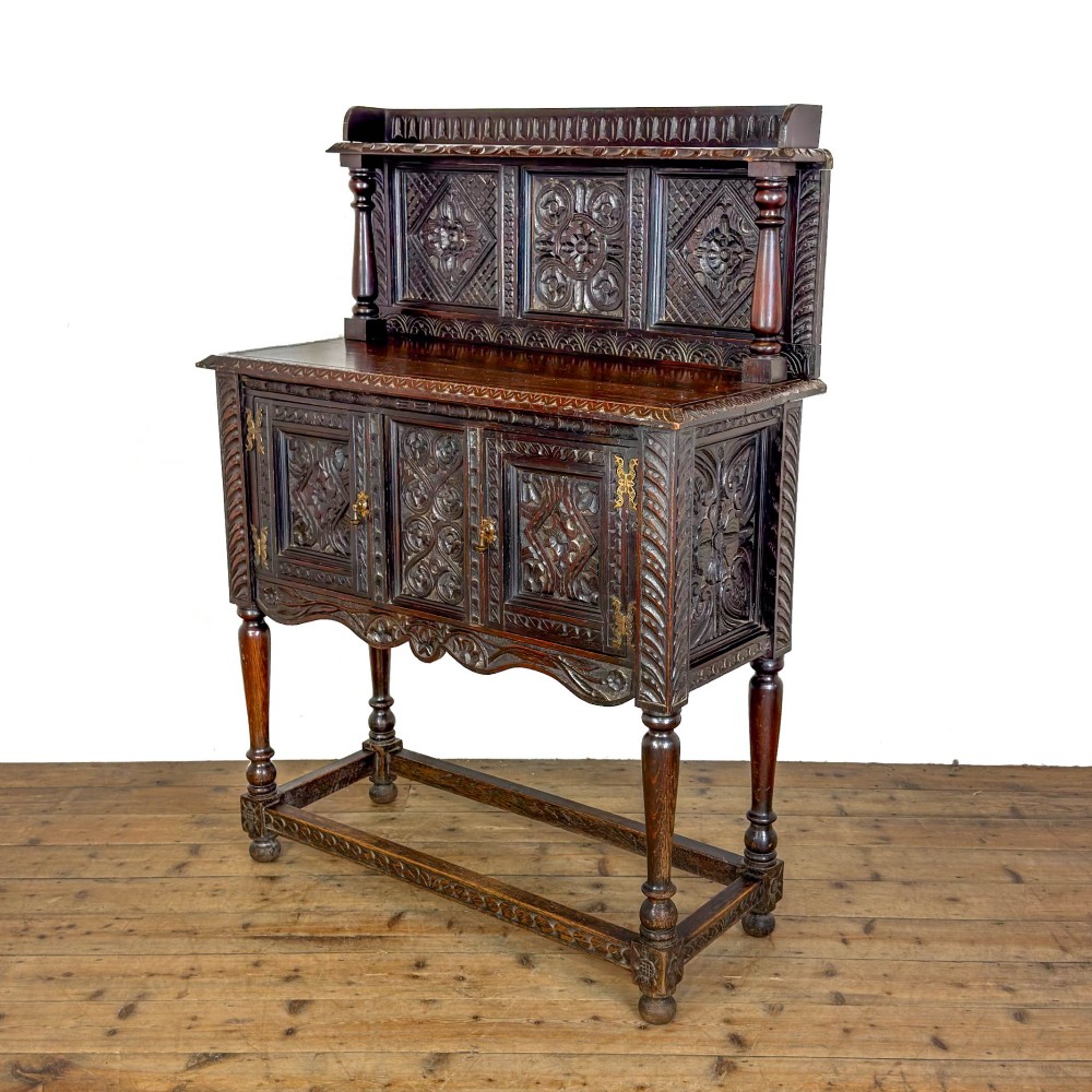 A dark carved oak Victorian sideboard with a tiered top shelf, detailed geometric carved door panels, brass hinges, and turned legs with a lower stretcher rail. View VI