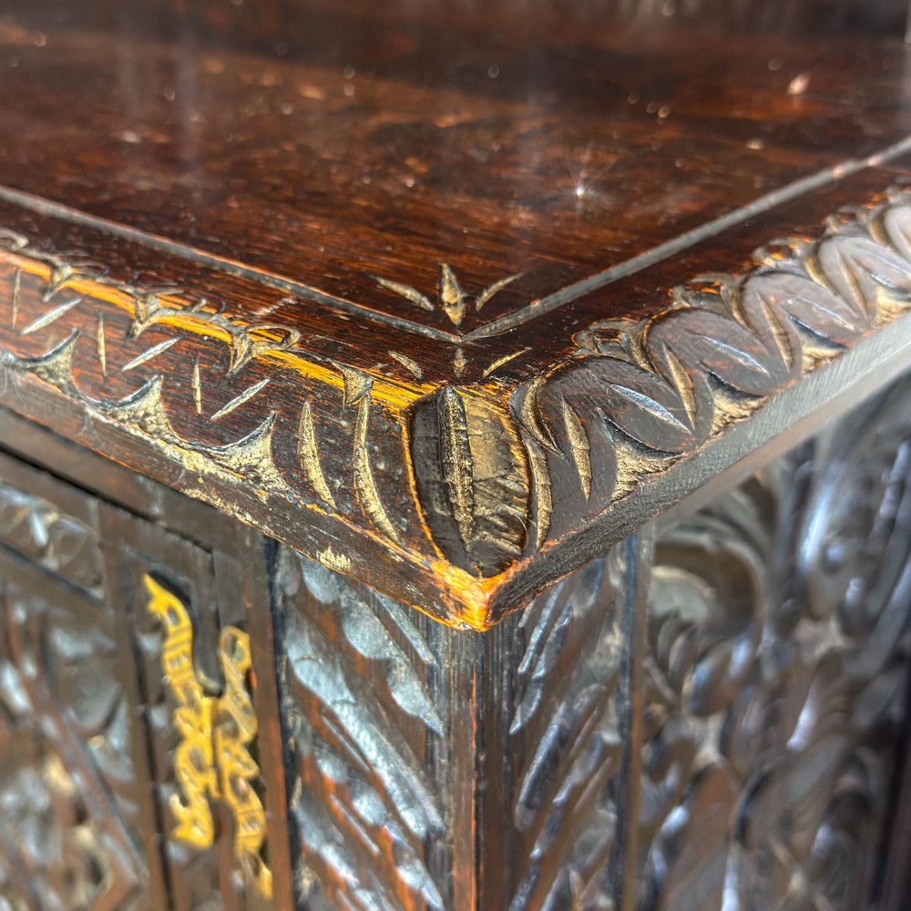 A dark carved oak Victorian sideboard with a tiered top shelf, detailed geometric carved door panels, brass hinges, and turned legs with a lower stretcher rail. View VII