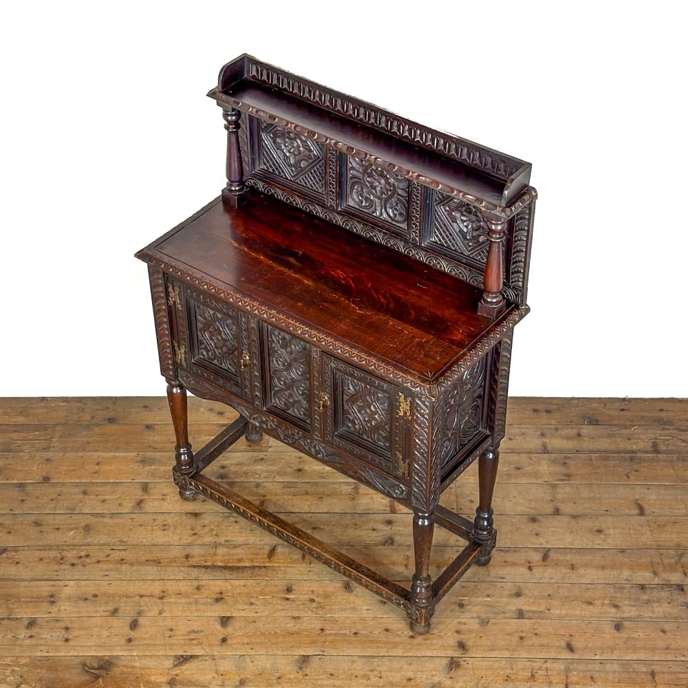 A dark carved oak Victorian sideboard with a tiered top shelf, detailed geometric carved door panels, brass hinges, and turned legs with a lower stretcher rail. View VIII