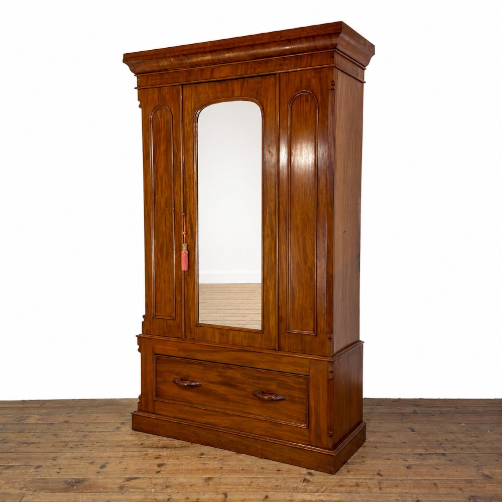 A tall Victorian mahogany wardrobe featuring an arched central mirrored door flanked by wooden side panels over a wide base drawer.