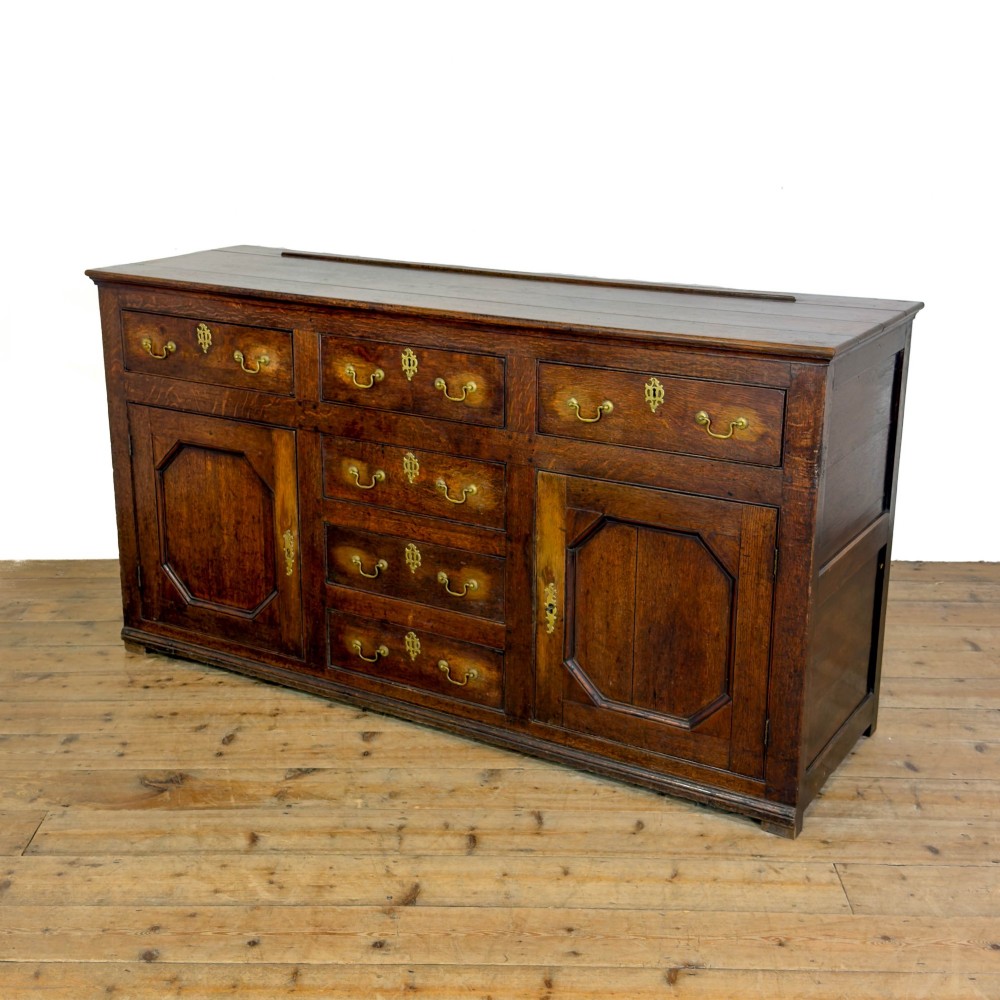 An early nineteenth-century dark oak dresser base featuring multiple drawers with brass handles, twin panelled cupboard doors, and a solid wood plinth base. View IV