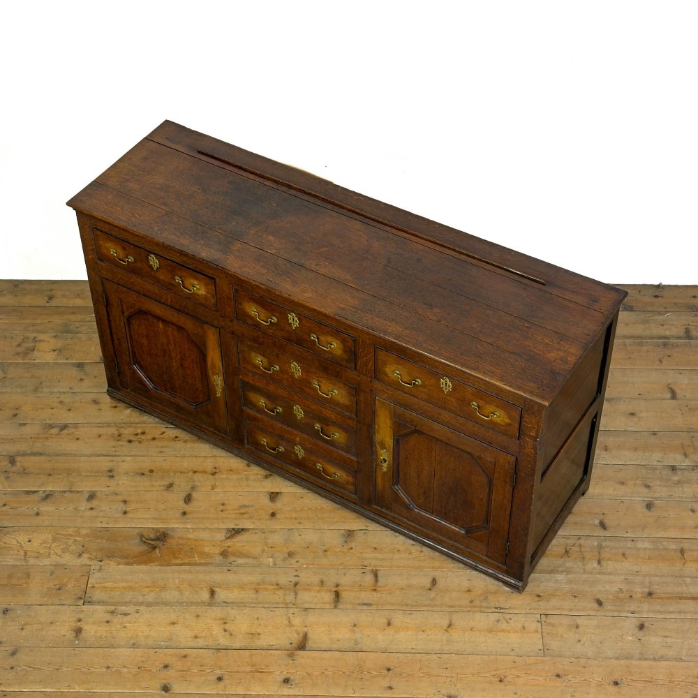 An early nineteenth-century dark oak dresser base featuring multiple drawers with brass handles, twin panelled cupboard doors, and a solid wood plinth base. View V