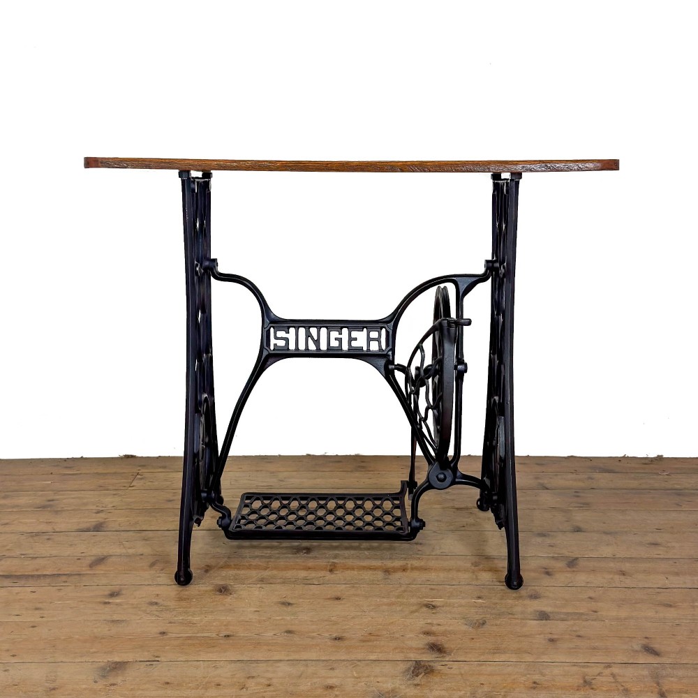 An antique black cast iron Singer treadle sewing machine base featuring a central brand crest and flywheel, fitted with a rectangular wooden tabletop.