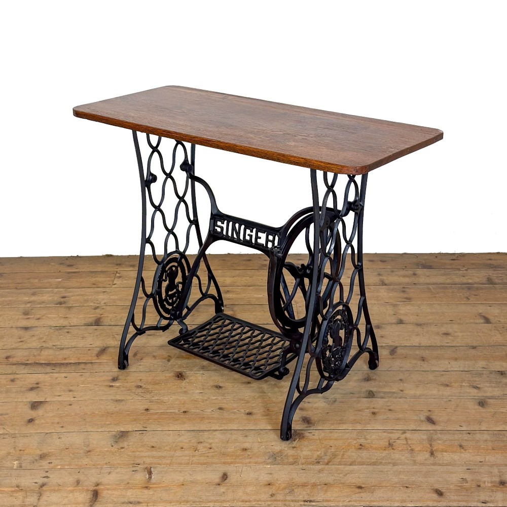 An antique black cast iron Singer treadle sewing machine base featuring a central brand crest and flywheel, fitted with a rectangular wooden tabletop. View II