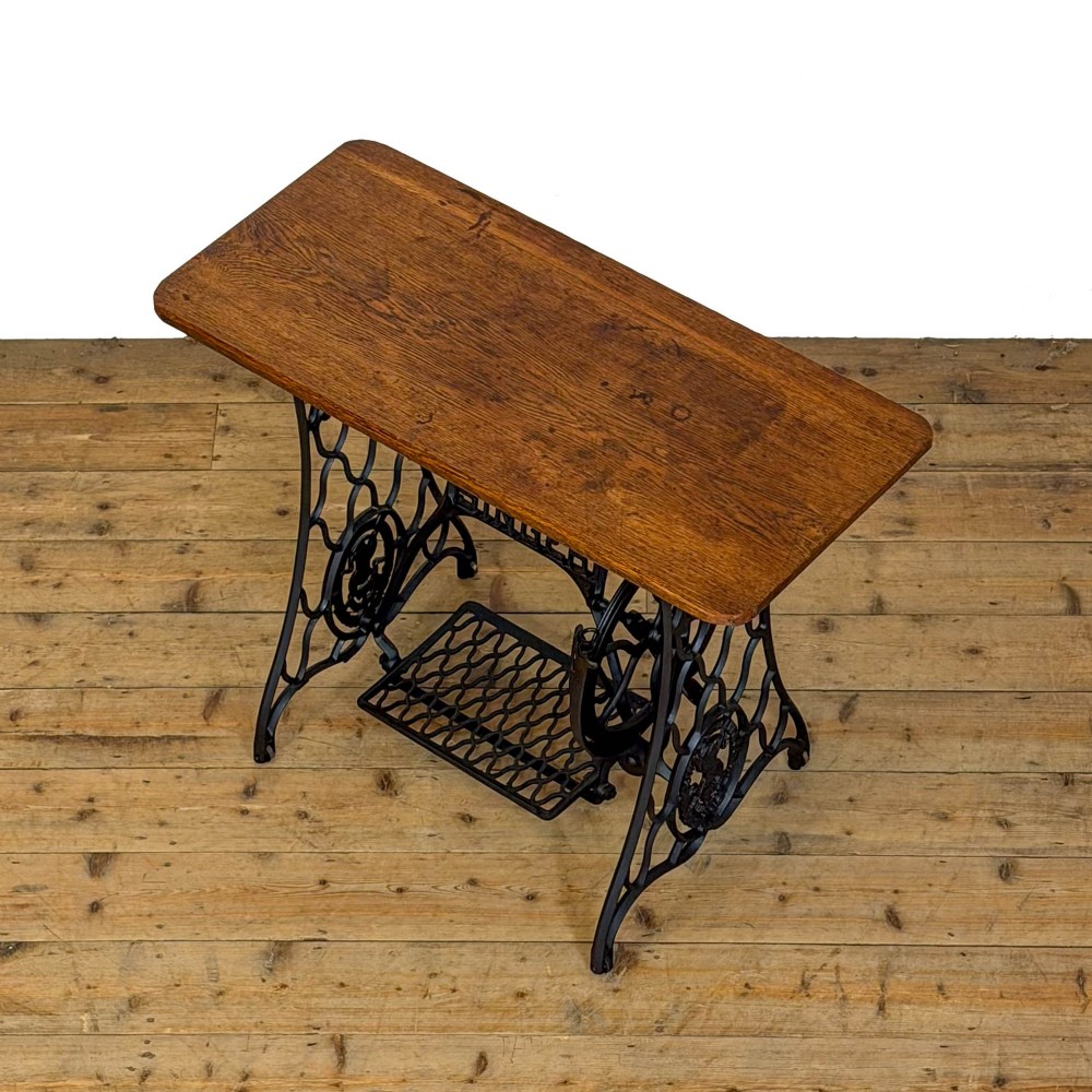 An antique black cast iron Singer treadle sewing machine base featuring a central brand crest and flywheel, fitted with a rectangular wooden tabletop. View III