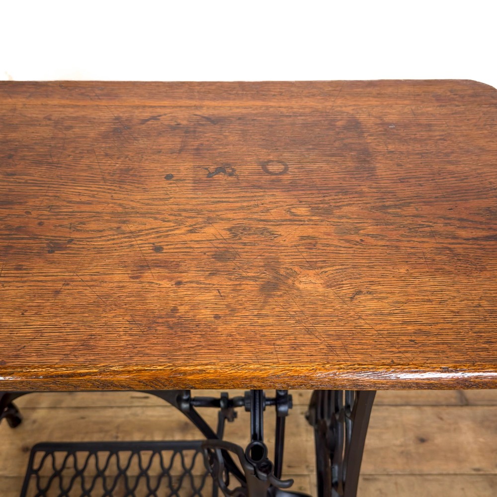 An antique black cast iron Singer treadle sewing machine base featuring a central brand crest and flywheel, fitted with a rectangular wooden tabletop. View IV