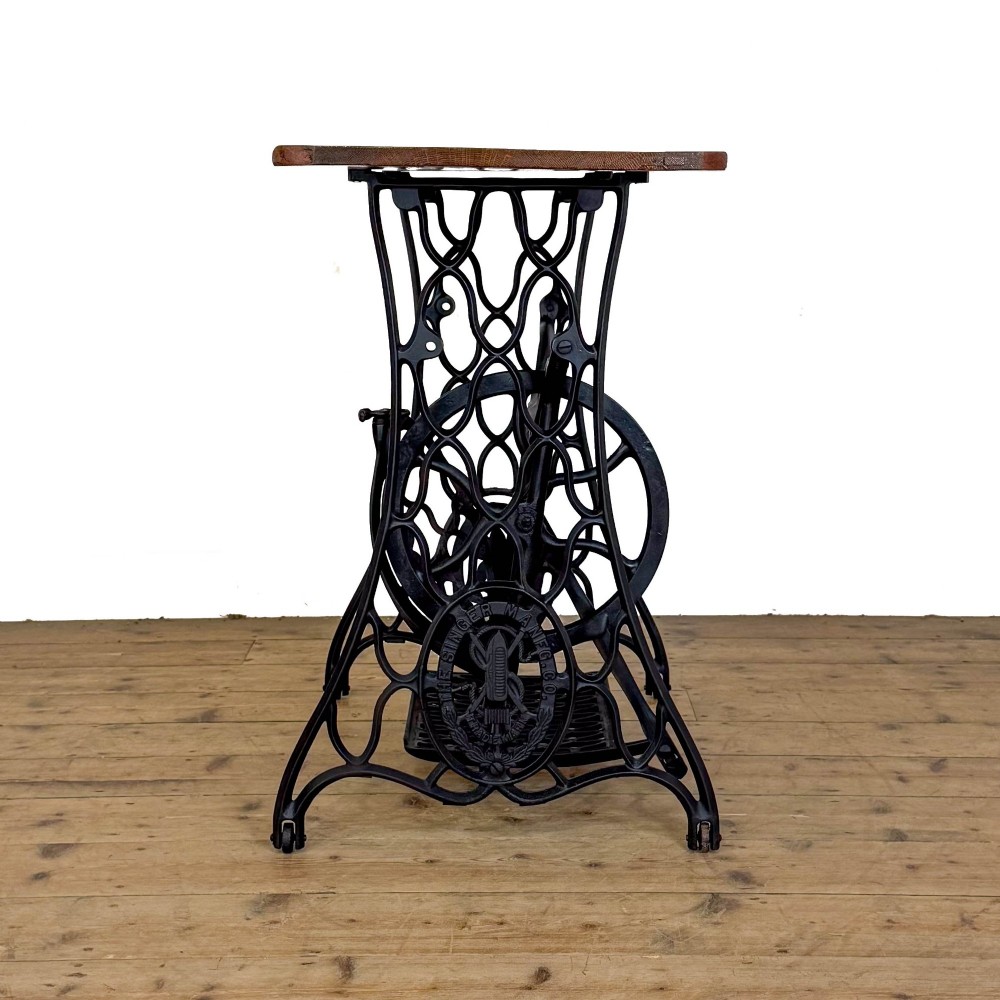 An antique black cast iron Singer treadle sewing machine base featuring a central brand crest and flywheel, fitted with a rectangular wooden tabletop. View V