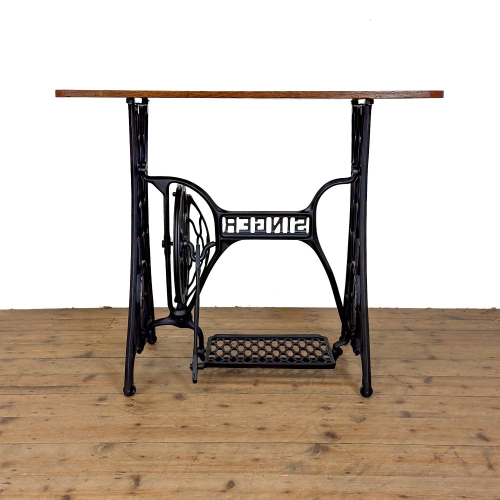 An antique black cast iron Singer treadle sewing machine base featuring a central brand crest and flywheel, fitted with a rectangular wooden tabletop. View VII