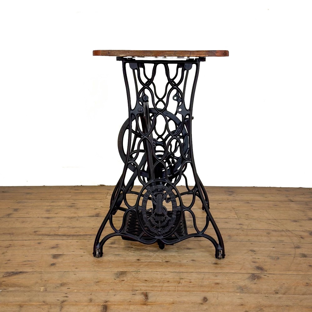 An antique black cast iron Singer treadle sewing machine base featuring a central brand crest and flywheel, fitted with a rectangular wooden tabletop. View VIII