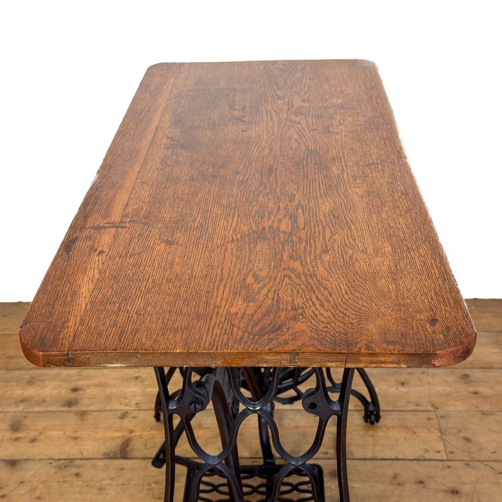 An antique black cast iron Singer treadle sewing machine base featuring a central brand crest and flywheel, fitted with a rectangular wooden tabletop. View IX