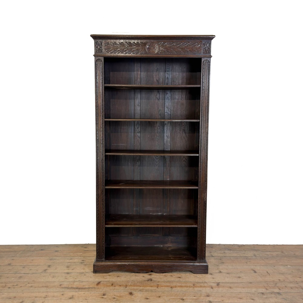 A tall dark oak open bookcase with multiple shelves, featuring carved floral scrollwork along the top frieze and fluted side columns.