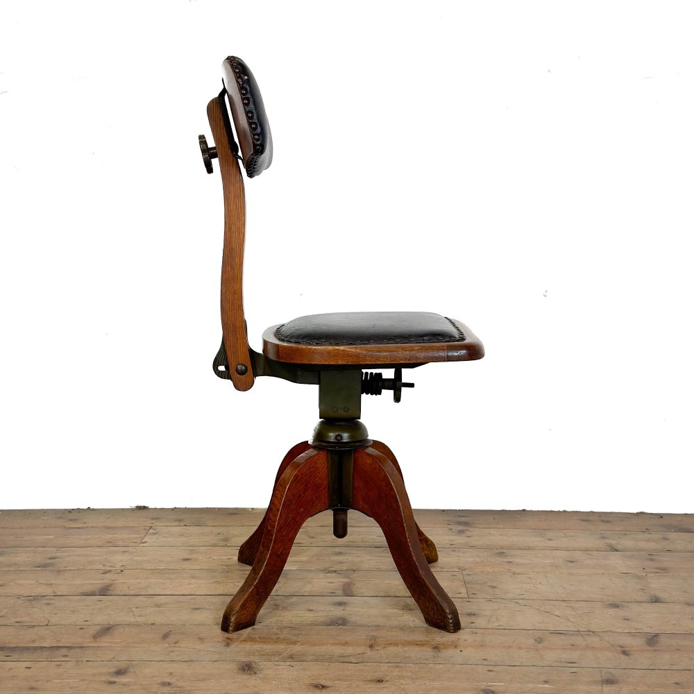 An early 20th-century oak swivel desk chair featuring dark studded leather upholstery, a curved lumbar backrest, and a four-star wooden base. View V