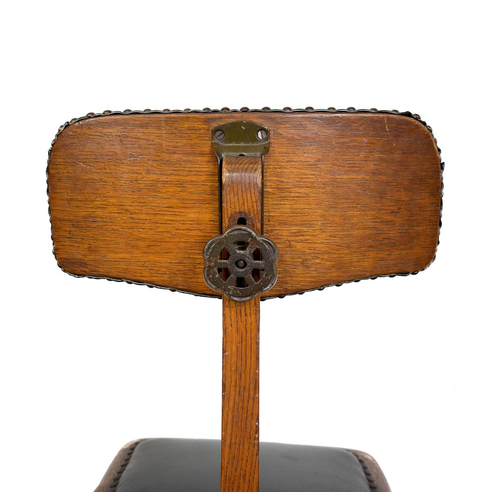 An early 20th-century oak swivel desk chair featuring dark studded leather upholstery, a curved lumbar backrest, and a four-star wooden base. View VI