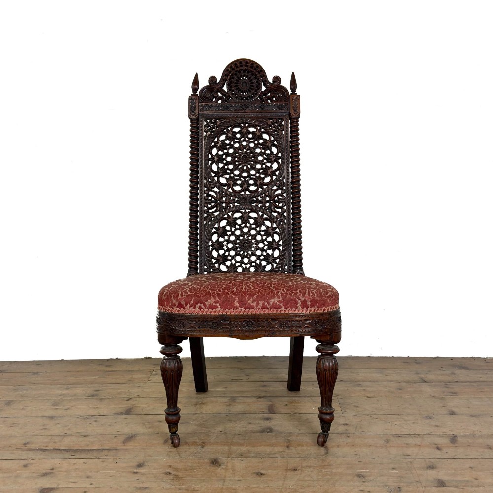 A 19th-century Anglo-Indian chair with an intricately hand-carved lattice backrest, dark wood frame, turned legs on castors, and a patterned red upholstered seat.