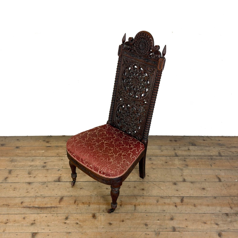 A 19th-century Anglo-Indian chair with an intricately hand-carved lattice backrest, dark wood frame, turned legs on castors, and a patterned red upholstered seat. View IV