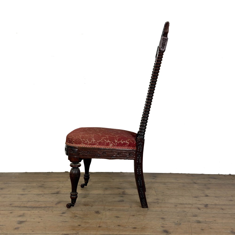 A 19th-century Anglo-Indian chair with an intricately hand-carved lattice backrest, dark wood frame, turned legs on castors, and a patterned red upholstered seat. View V