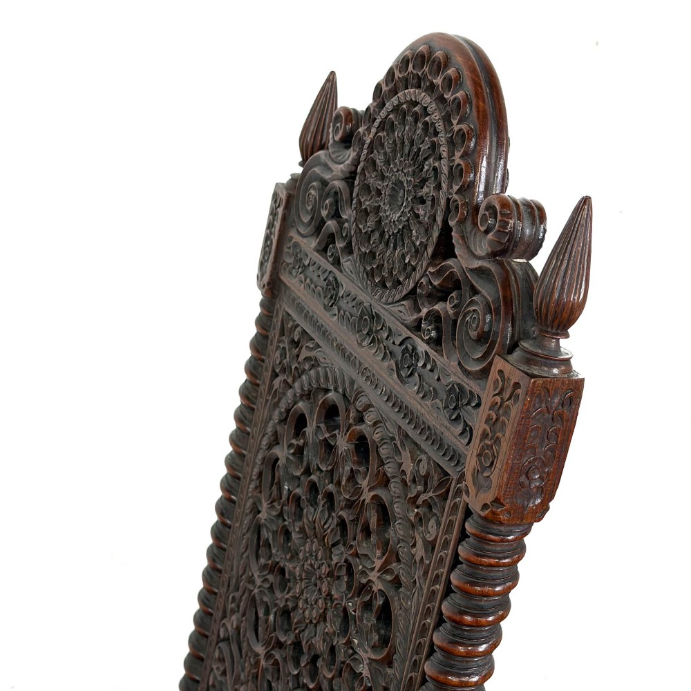 A 19th-century Anglo-Indian chair with an intricately hand-carved lattice backrest, dark wood frame, turned legs on castors, and a patterned red upholstered seat. View VI