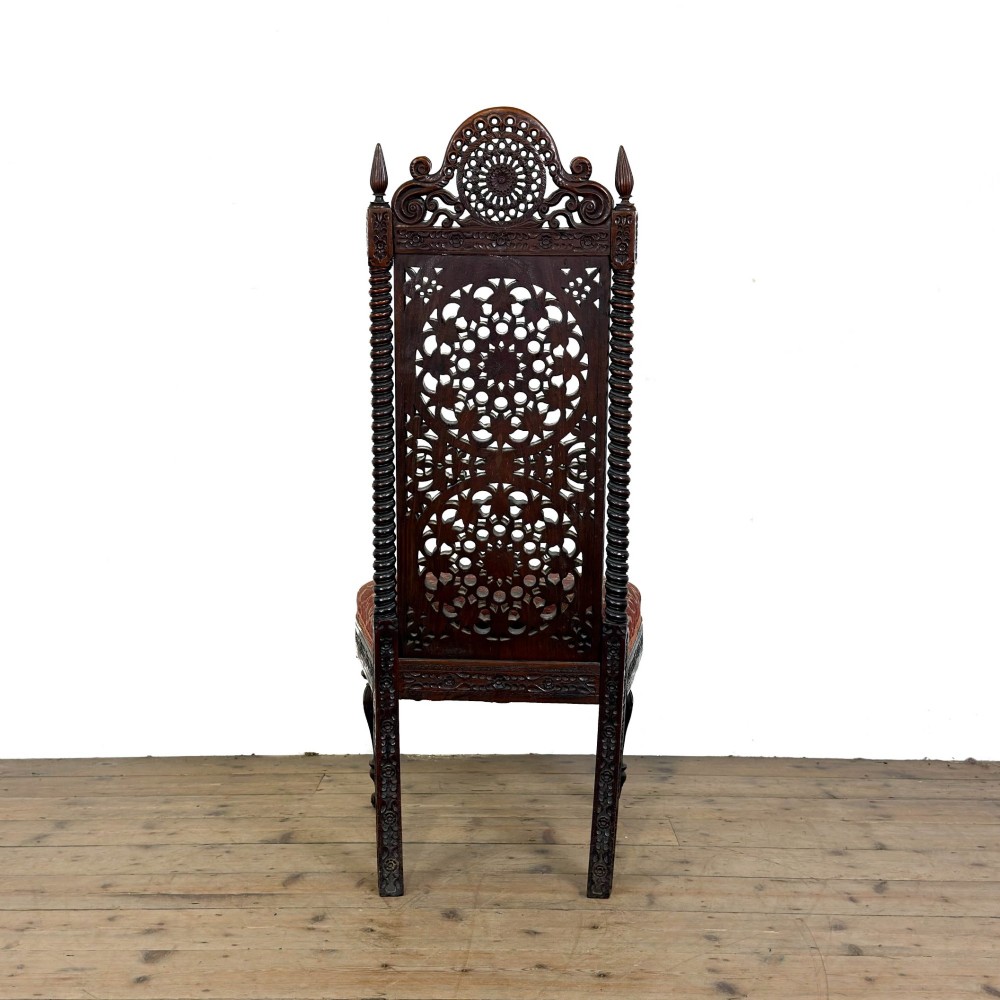 A 19th-century Anglo-Indian chair with an intricately hand-carved lattice backrest, dark wood frame, turned legs on castors, and a patterned red upholstered seat. View VII