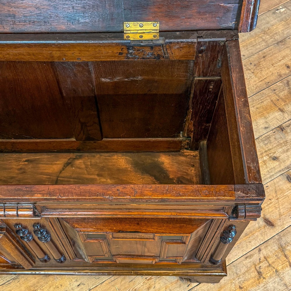A seventeenth-century style carved oak coffer featuring geometric panelling, split baluster turnings, and dark square inlay details on a plinth base. View V