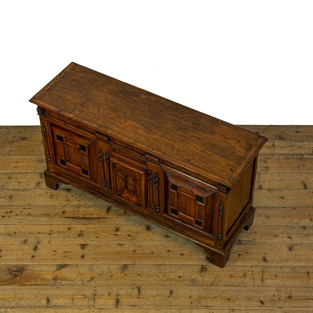 A seventeenth-century style carved oak coffer featuring geometric panelling, split baluster turnings, and dark square inlay details on a plinth base. View VII