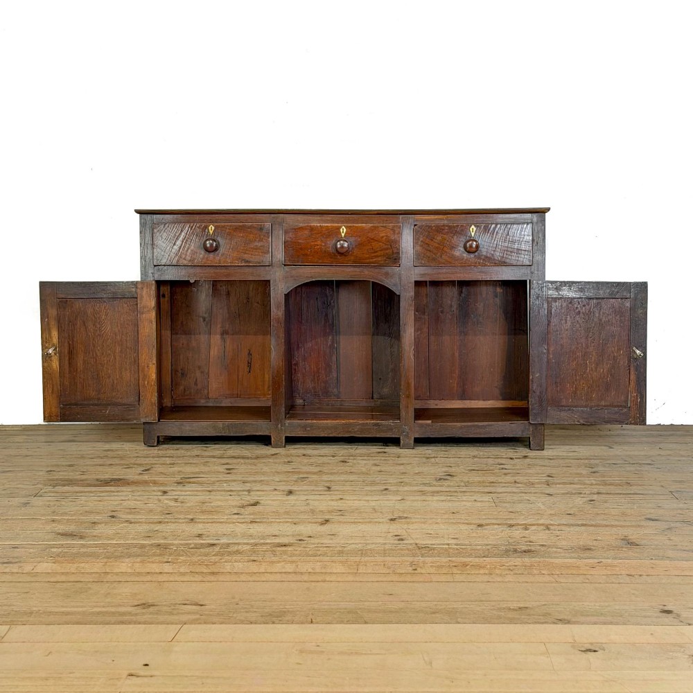 A dark oak antique dresser base featuring three top drawers with wooden knobs and brass keyholes above two panelled cupboards flanking a central open alcove. View II