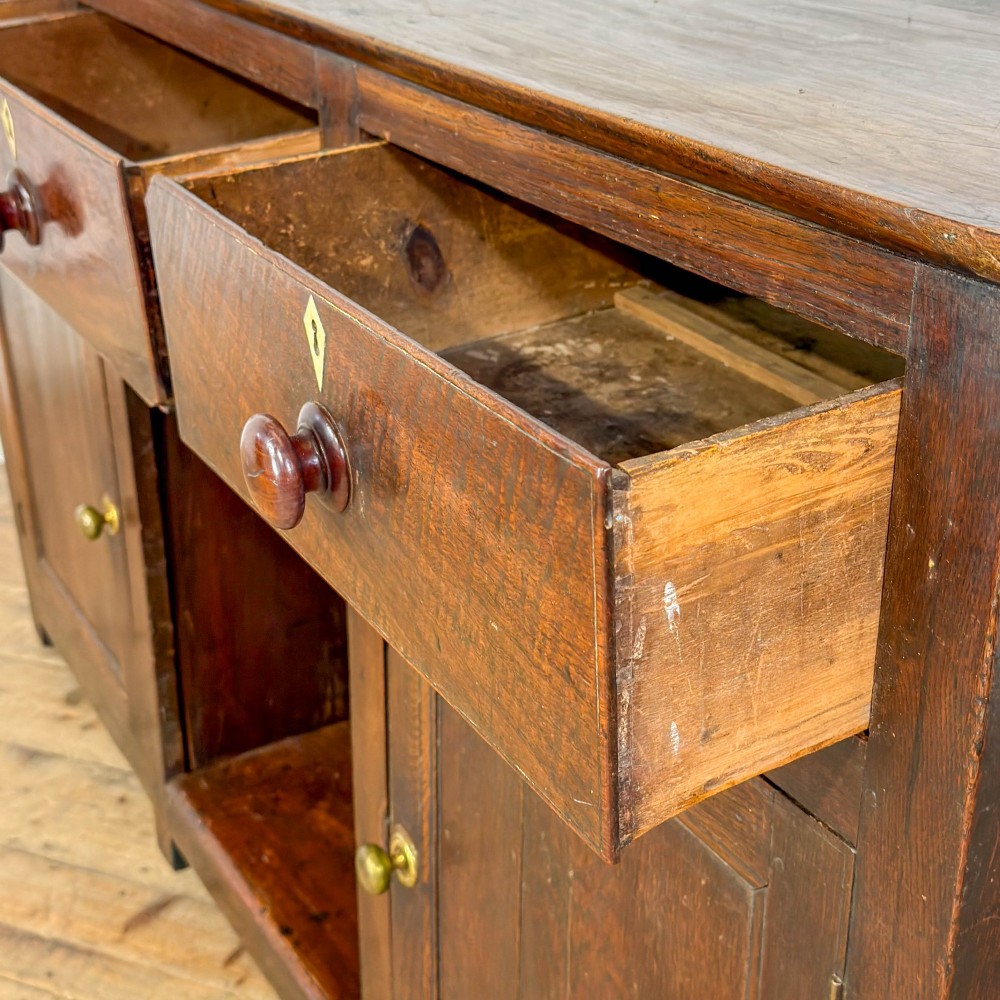 A dark oak antique dresser base featuring three top drawers with wooden knobs and brass keyholes above two panelled cupboards flanking a central open alcove. View V