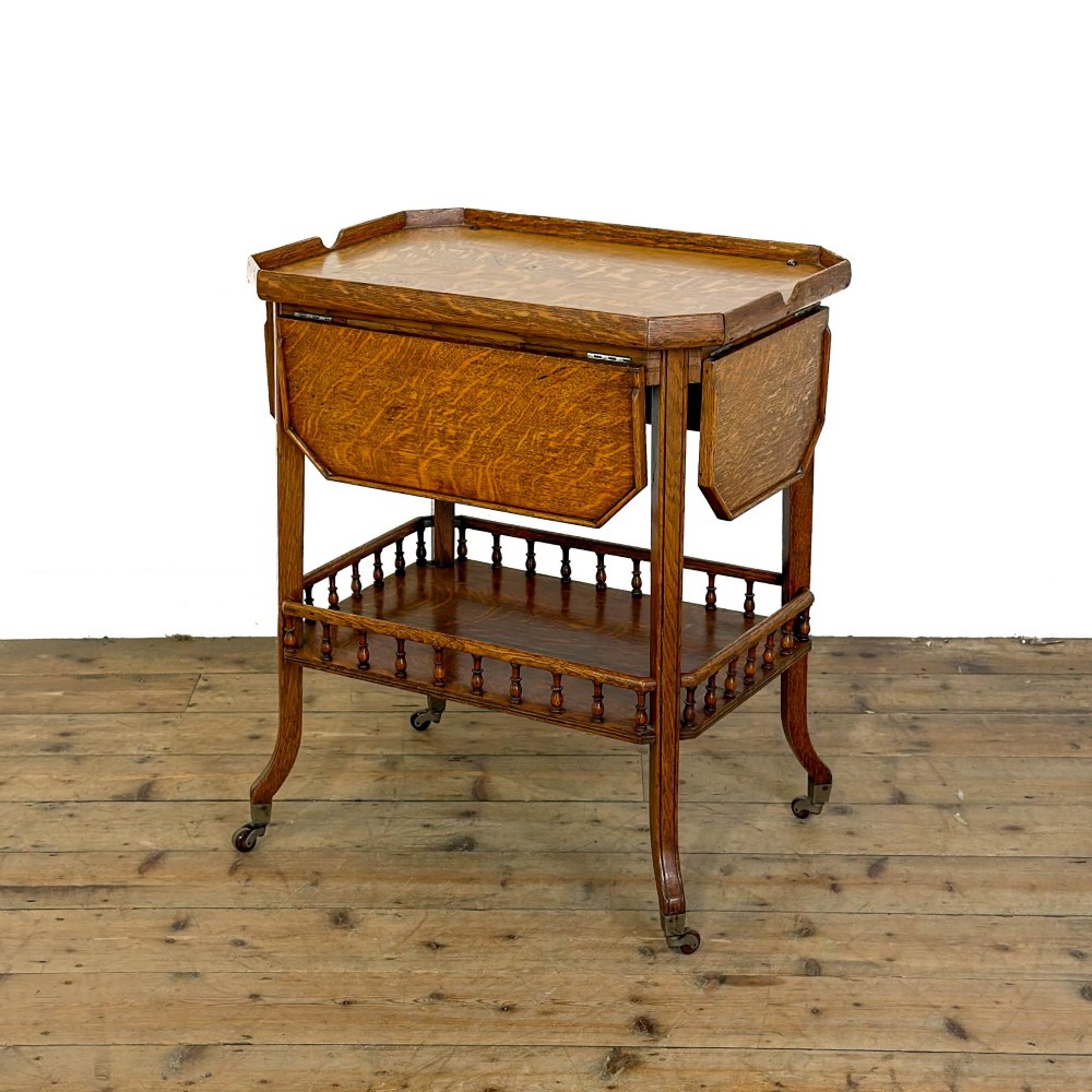 An Edwardian medium oak two-tier tea trolley featuring drop-leaf side panels, a spindle-turned gallery on the lower shelf, and brass castors.