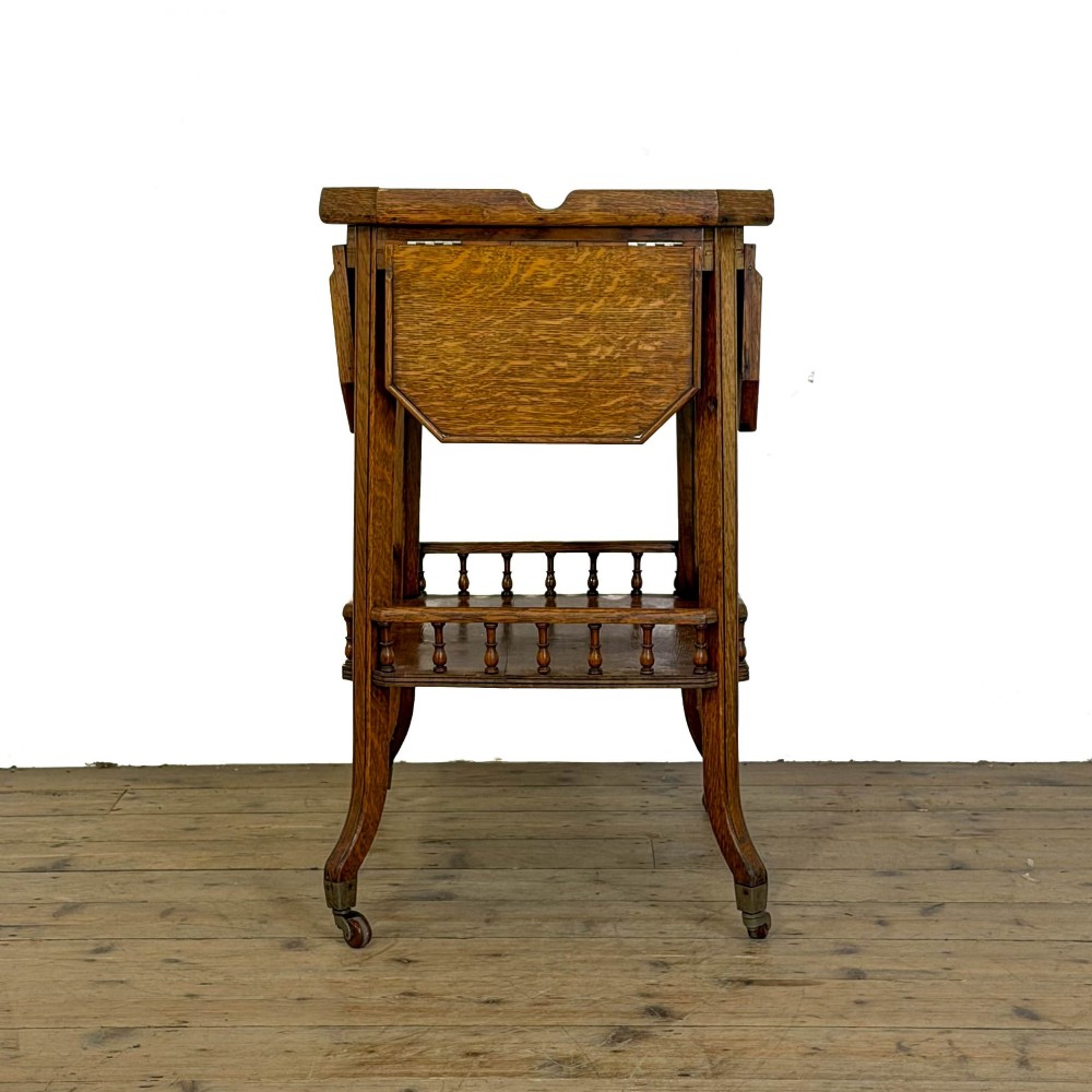 An Edwardian medium oak two-tier tea trolley featuring drop-leaf side panels, a spindle-turned gallery on the lower shelf, and brass castors. View II