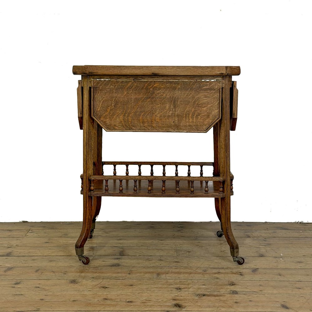 An Edwardian medium oak two-tier tea trolley featuring drop-leaf side panels, a spindle-turned gallery on the lower shelf, and brass castors. View III