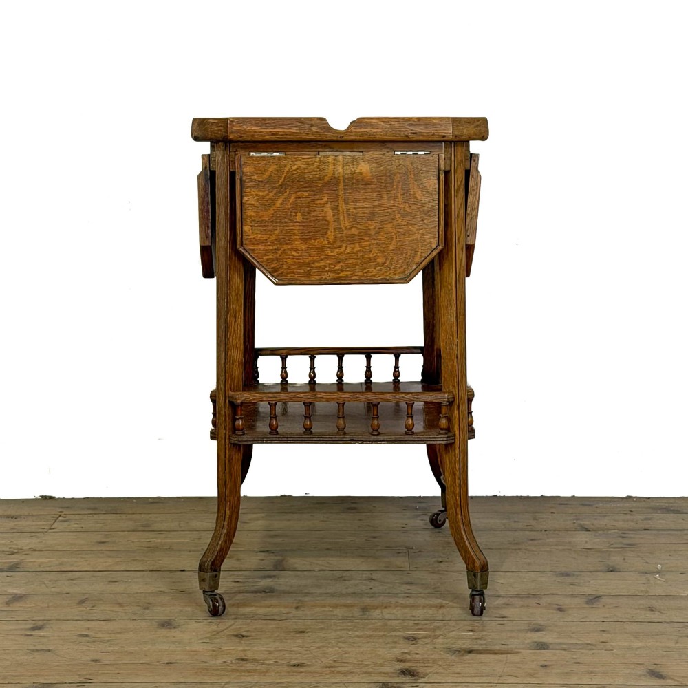 An Edwardian medium oak two-tier tea trolley featuring drop-leaf side panels, a spindle-turned gallery on the lower shelf, and brass castors. View IV