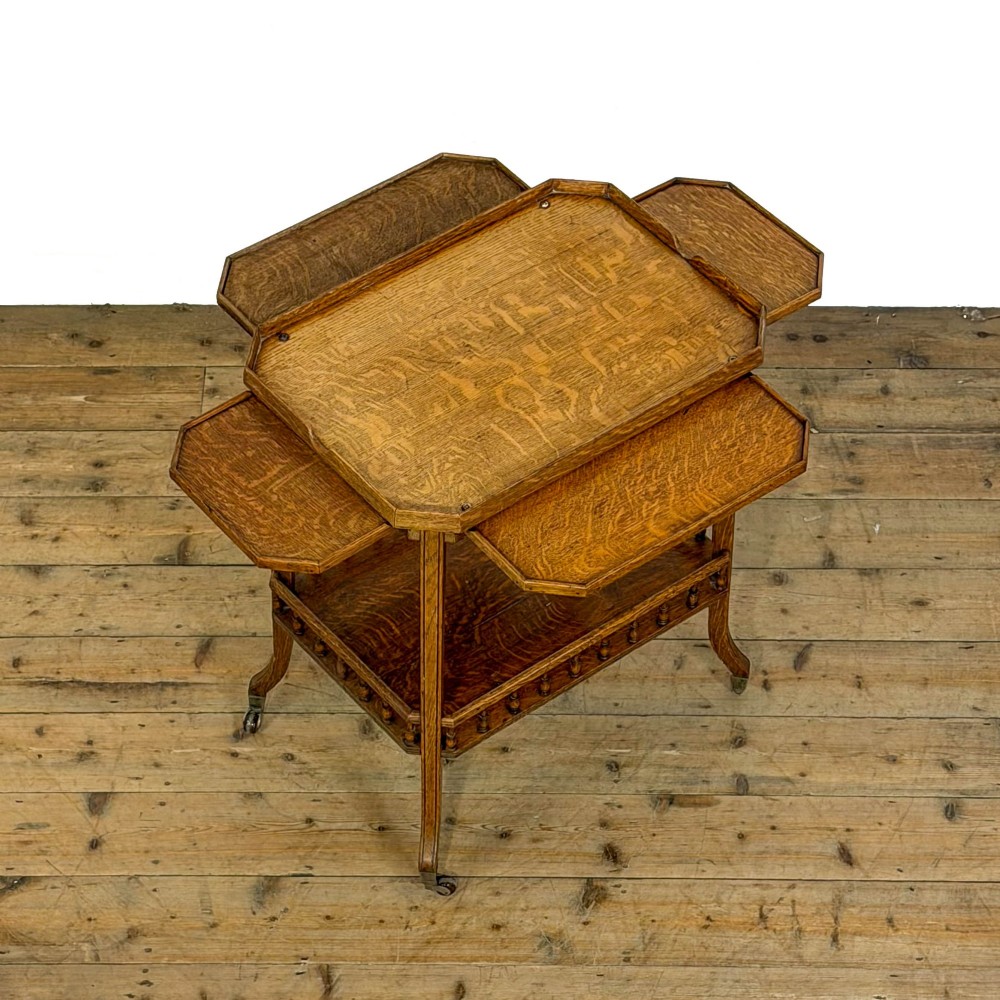 An Edwardian medium oak two-tier tea trolley featuring drop-leaf side panels, a spindle-turned gallery on the lower shelf, and brass castors. View V